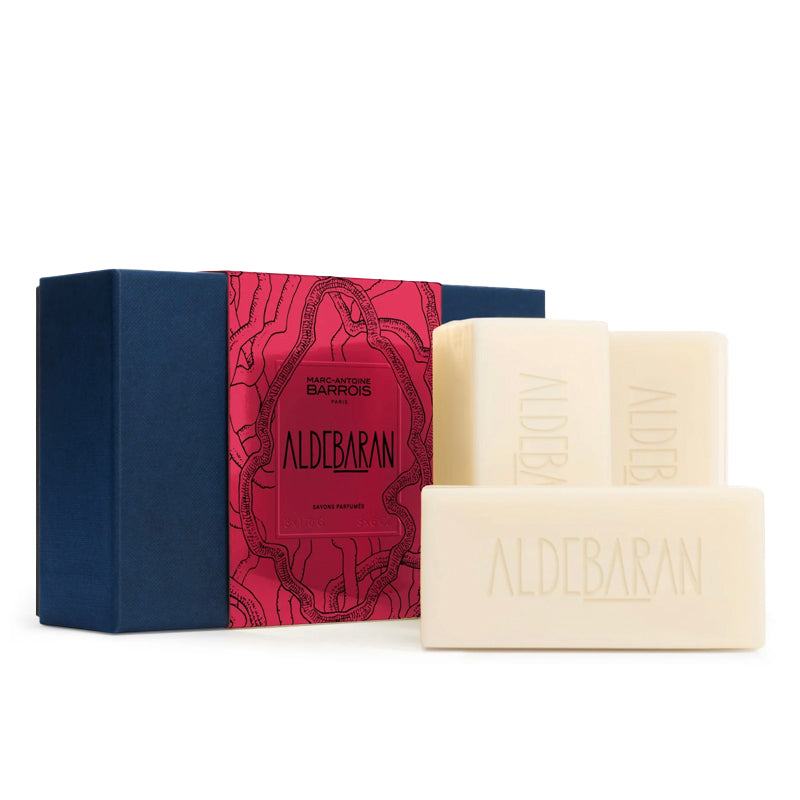 Three cream-colored ALDEBARAN soap bars displayed in front of their packaging box, which features a navy blue side and a vibrant pink patterned front panel.