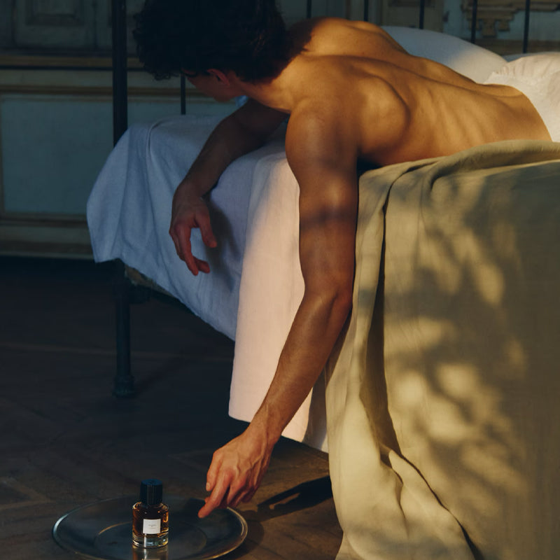 Man sitting on a bed with a bottle of Carmen perfume by Trudon on a tray in a dimly lit room.