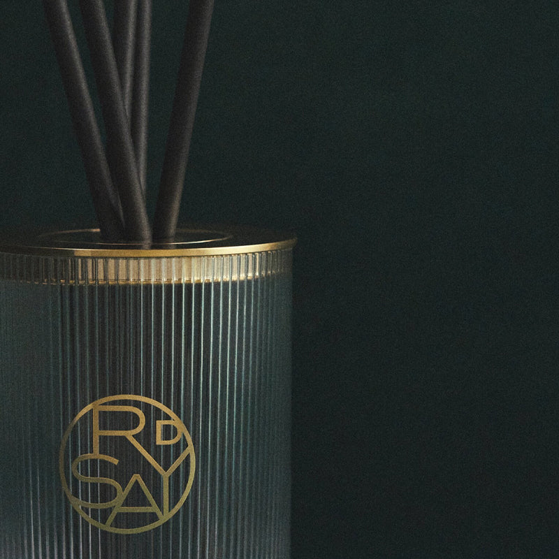 Decorative diffuser with black reeds and gold logo on a dark background
