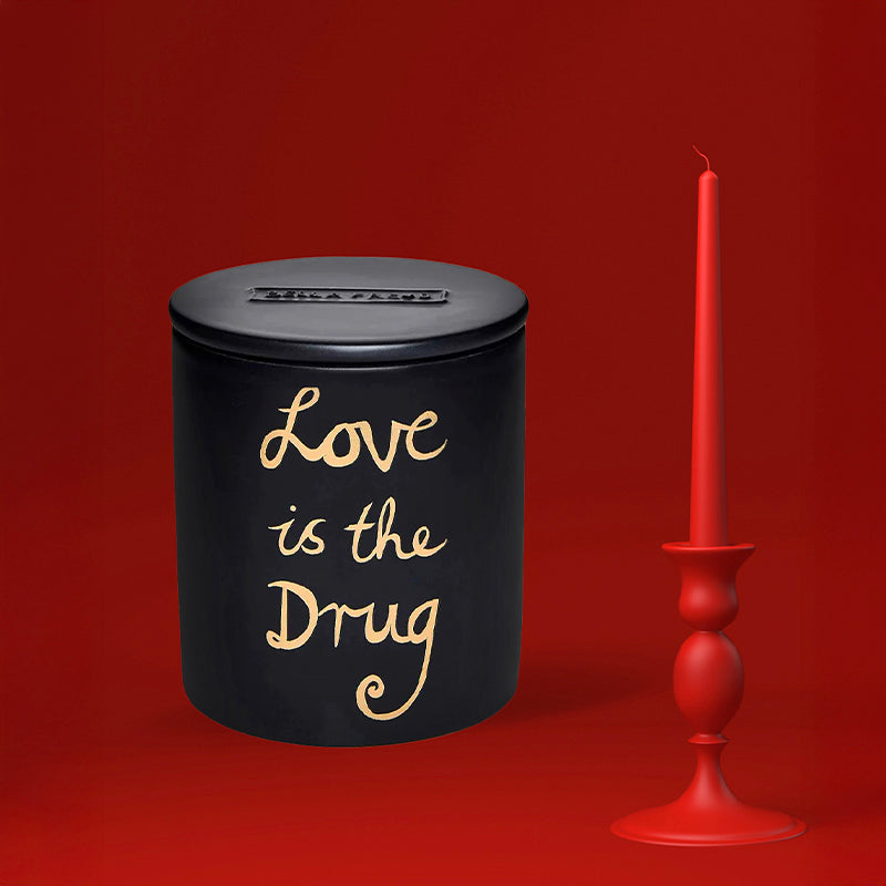 Black container with 'Love is the Drug' candle by Bella Freud. Shown with a red candlestick.