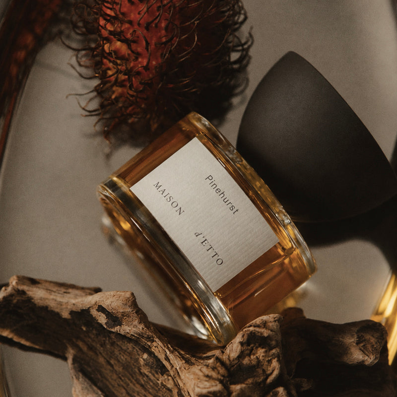 A Maison d'Etto Pinehurst flacon with green cap resting on a piece of drift wood and a lichee fruit