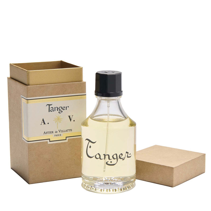 Tanger Parfum 3.4oz or 100ml bottle by Astier de Villatte. Clear bottle with the writing "Tanger" containing the clear golden fragrance. The cap is black. Next to it a brown paper box with the label that reads Tanger Astier de Villatte Paris and the initials A.V. with a golden palm tree symbol between the A. and V.