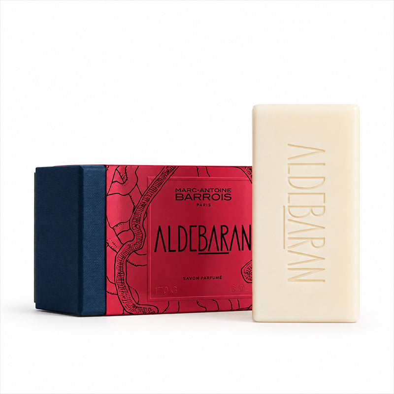 A bar of Marc-Antoine Barois Aldebaran perfume soap in an off-white color, standing next to its red and navy blue illustrated packaging box on a clean white background.