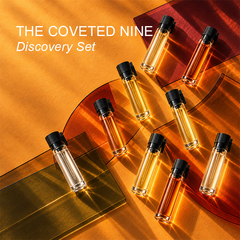 Nine black-capped fragrance sample vials in clear, golden, and amber tones arranged on an amber surface with translucent autumn-colored acrylic shapes.