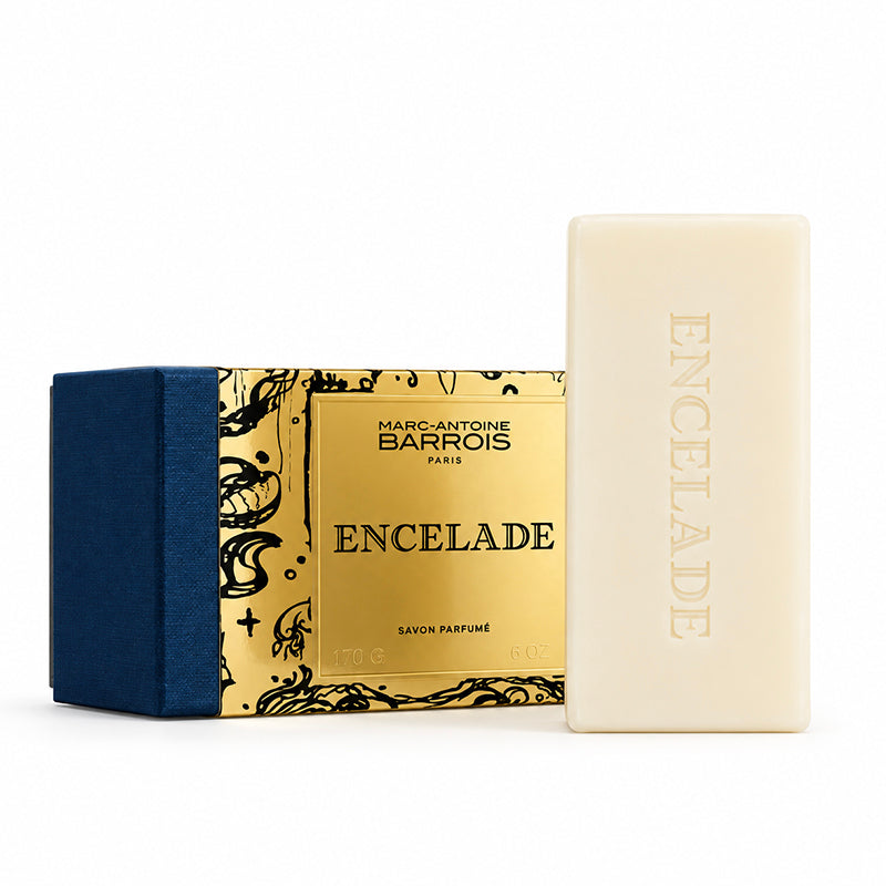 A bar of Marc-Antoine Barrois Encelade perfume soap next to its decorative packaging box, featuring a navy blue velvet side and a gold patterned front panel.