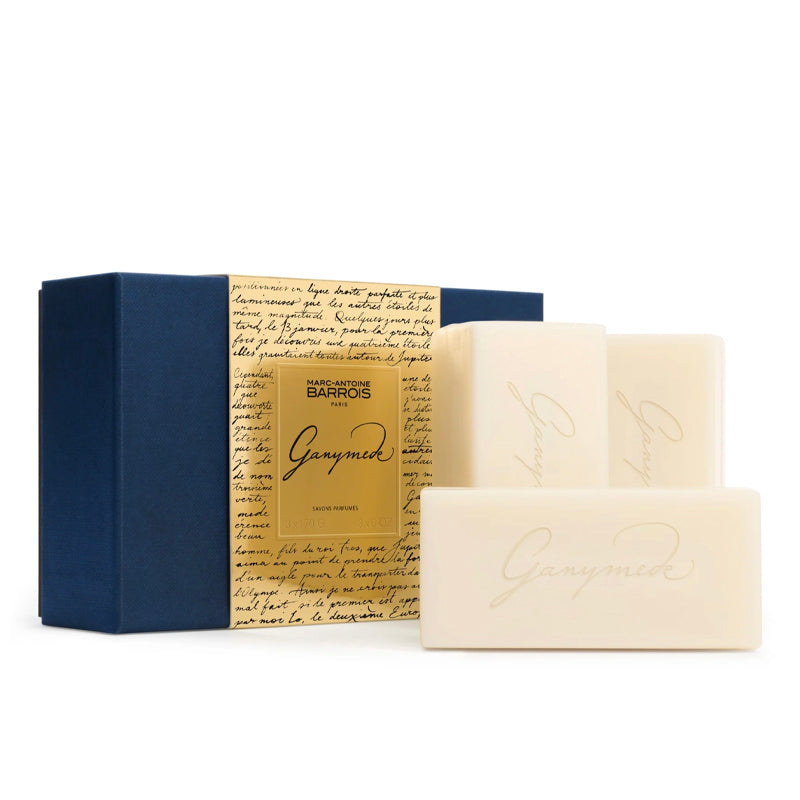 Three cream-colored rectangular bars of soap with elegant gold cursive writing, displayed in front of their packaging. The packaging consists of a navy blue box and a central gold label with cursive writing, all on a clean white background.