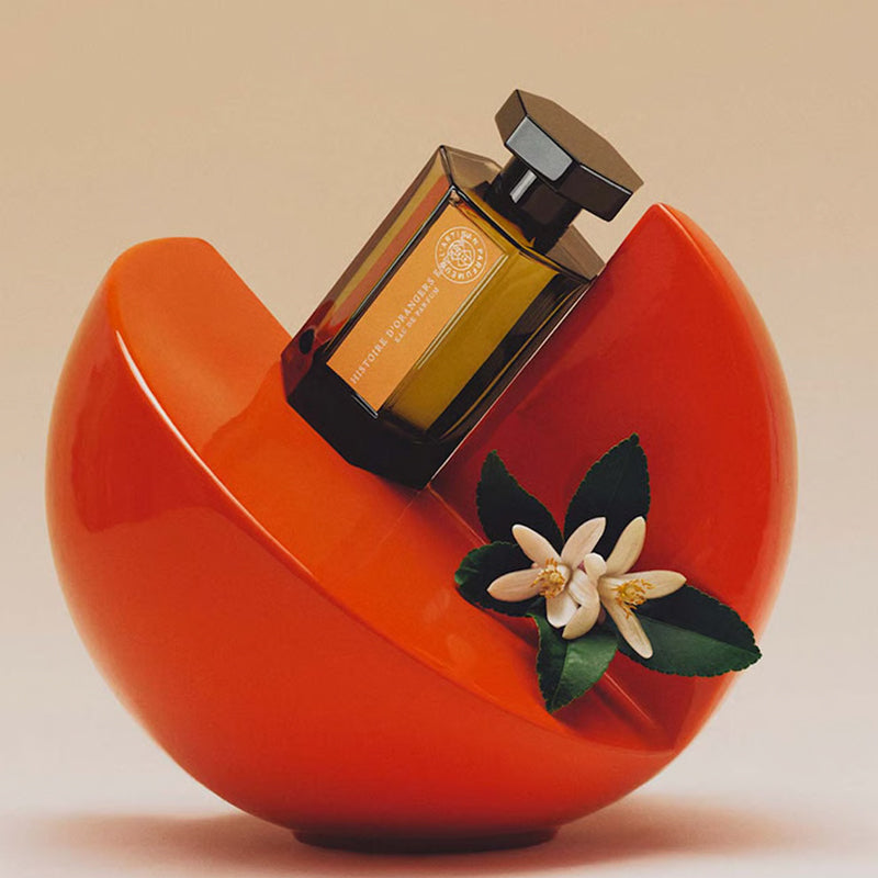 Perfume bottle on a red heart-shaped stand with a flower decoration against a beige background. Histoire d'Oranger by L'Artisan Parfumeur