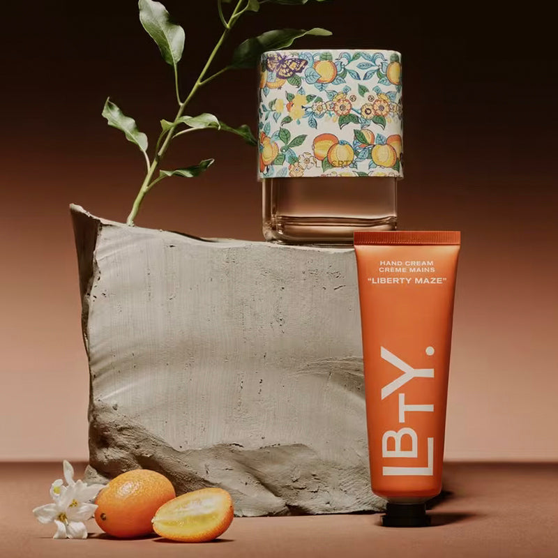 Hand cream, floral-patterned bottle, and oranges on a textured surface with a brown background. Liberty Maze - Hand Cream | Liberty Beauty | AEDES.COM