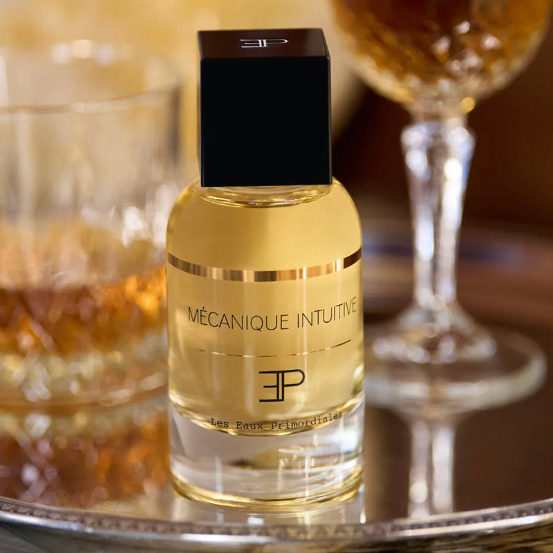 Perfume bottle labeled 'Mécanique Intuitive' on a reflective surface with blurred glasses in the background. Mécanique Intuitive - EdP 100ml Superclassique Collection | Les Eaux Primordiales