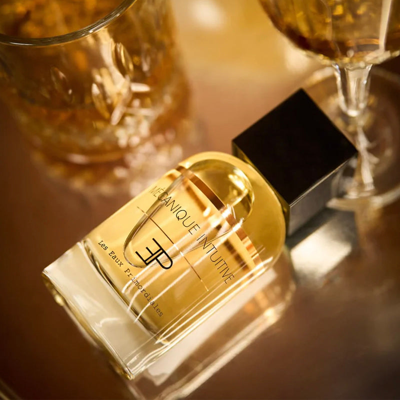 Perfume bottle with gold cap and black base on a reflective surface with blurred glasses in the background. Mécanique Intuitive - EdP 100ml Superclassique Collection | Les Eaux Primordiales