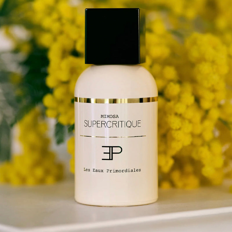 White bottle with black cap labeled 'Supercritique' against a yellow floral background. Mimosa EdP - Supercritique Collection | LES EAUX PRIMORDIALES